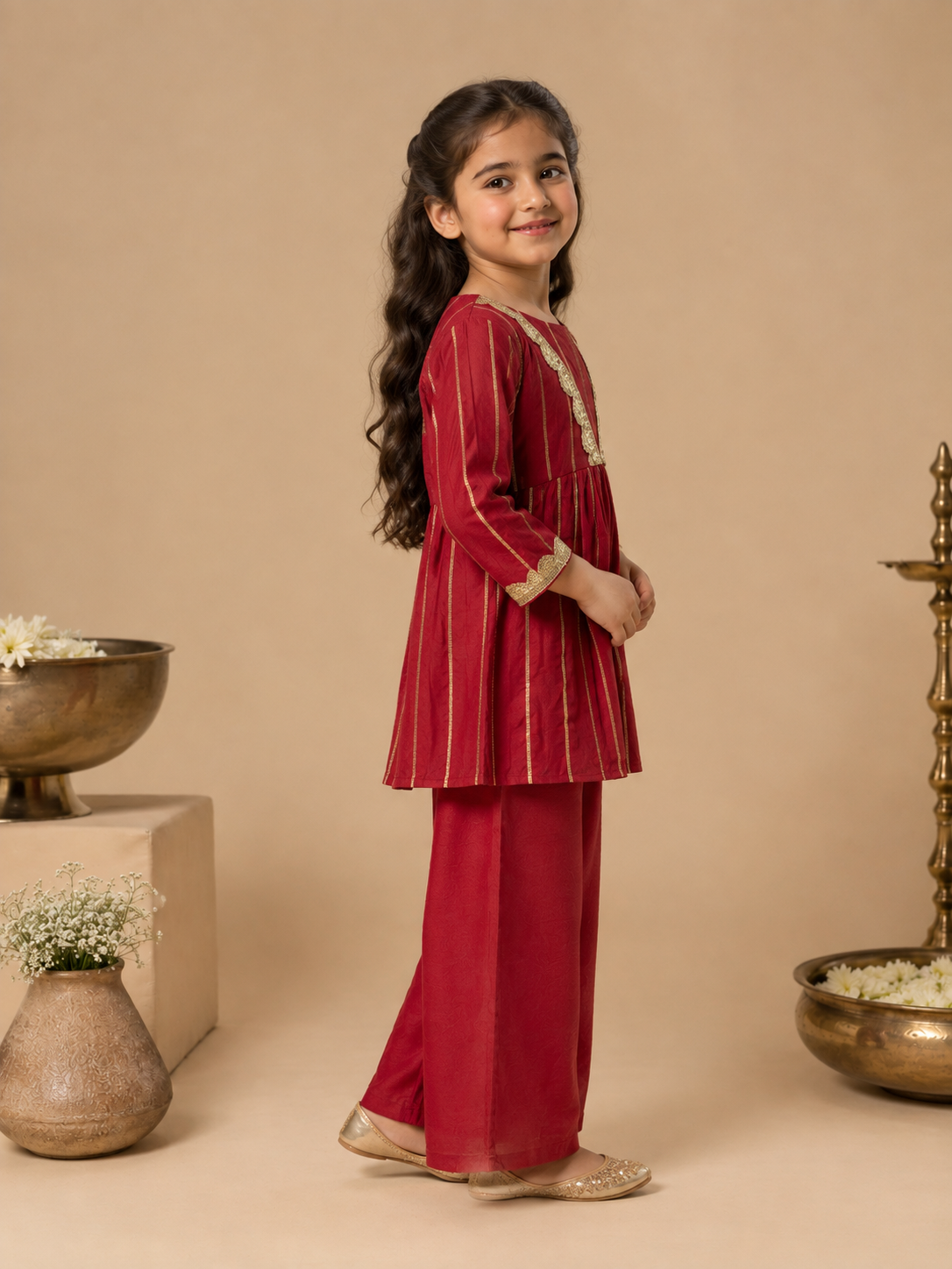 A young girl modeling a traditional Indian outfit consisting of a deep red kurta with gold vertical stripes and matching wide-leg trousers, set against a warm beige background with decorative props.