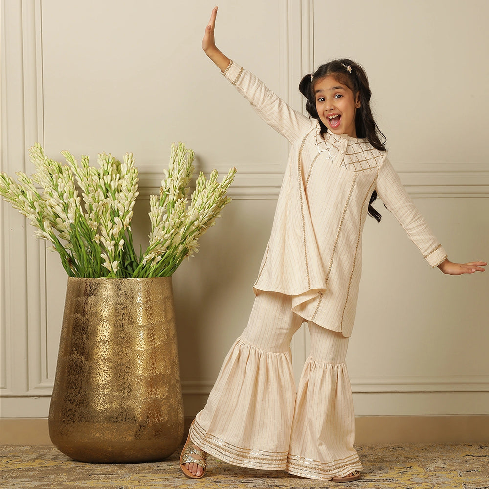 
                  
                    Elegant in its simplicity, the Rajni Sharara Set is a refined ensemble for special occasions. Crafted in a soft ivory-peach hue with subtle&nbsp;pinstripes, this set features a straight-cut kurta with delicate gold threadwork detailing on the yoke and sleeves. 
                  
                